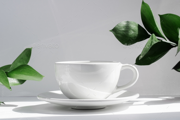 White tea or coffee mug and saucer, on a white table in the rays of the sun