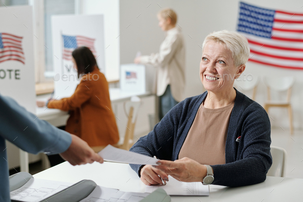 Happy mature woman passing ballot paper to young voter at polling place - Stock Photo - Images