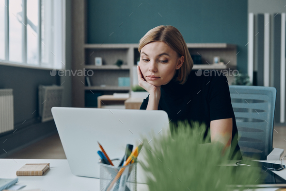 Bored young businesswoman looking at laptop while sitting at the office desk - Stock Photo - Images