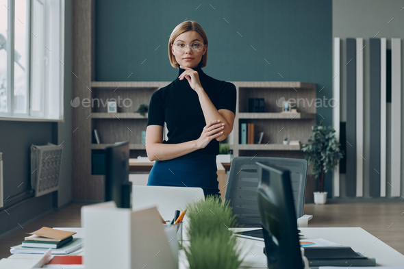 Serious businesswoman looking at camera while standing near her working place in office - Stock Photo - Images