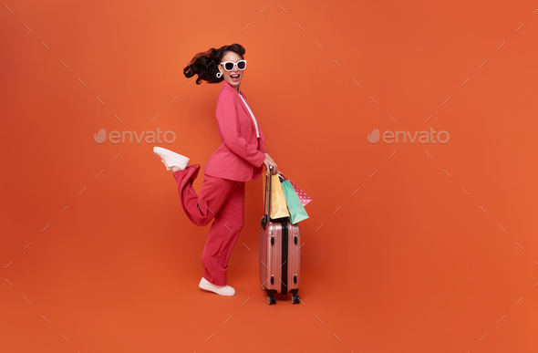 Happy young woman traveler drag luggage and shopping bag isolated on orange copy space background