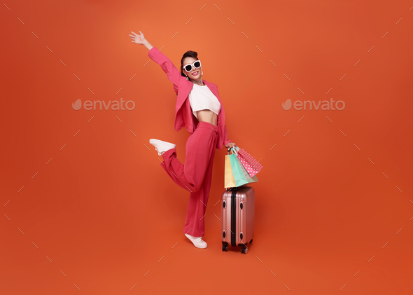 Happy young woman traveler drag luggage and shopping bag isolated on orange copy space background