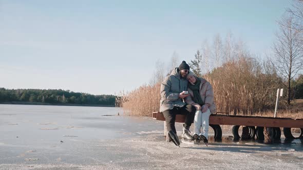 Cute Couple Sit on a Lake Dock in Winter Drink Hot Tea From Thermos and Hug