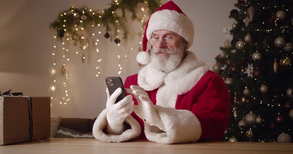 Santa Claus Using Smartphone Sitting Against a Lit Christmas Tree Background Smiles Laughs Rejoices alt