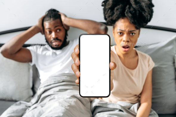 Shocked puzzled african american couple, sit in a cozy bed in the bedroom, woman shows smartphone
