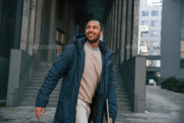 In blue jacket and with laptop in hands. Handsome black man is outdoors near the business building - Stock Photo - Images