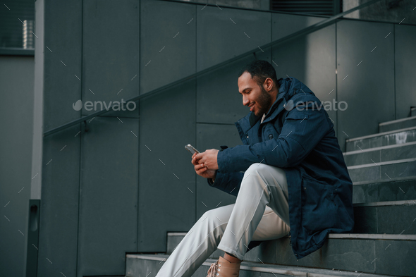 on the stairs with smartphone in hands. Handsome black man is outdoors near the business building - Stock Photo - Images