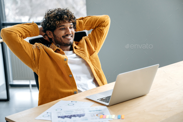 Positive satisfied Indian man, in stylish casual clothes, company employee, sits at his work desk