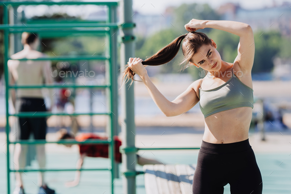 Tired sporty brunette girl at sport court in bra and leggings making  ponytail looks at camera. Stock Photo by ionadidishvili