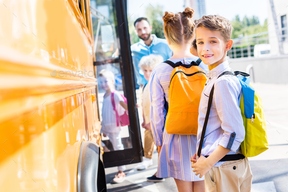 adorable schoolboy entering school bus with classmates while teacher standing near door