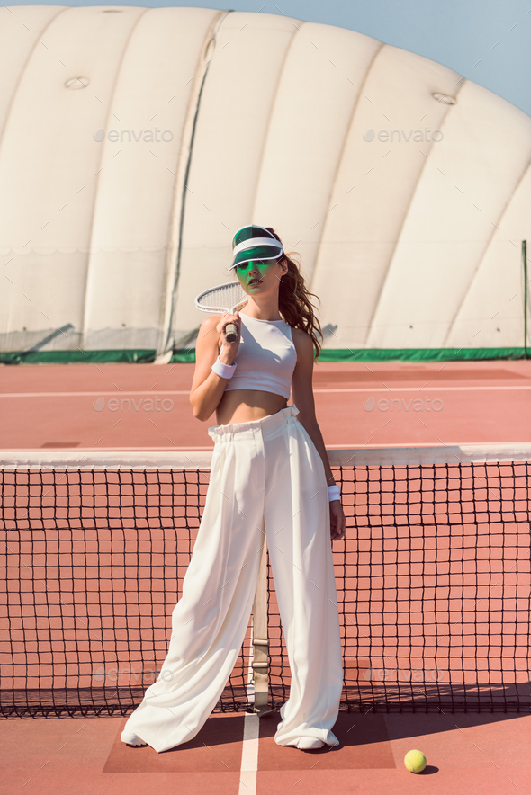 stylish woman in white clothing and cap with tennis racket posing at tennis net on court