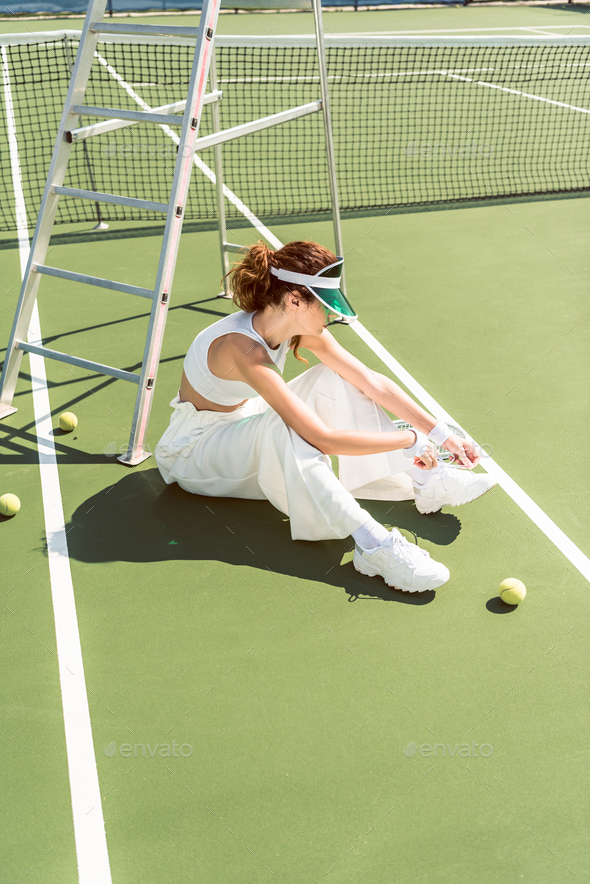 side view of young woman in stylish white clothing and cap tying shoelaces on tennis court with