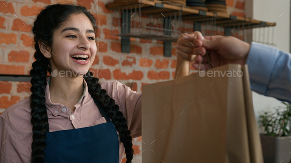 Friendly young indian woman waitress barista giving takeaway food bag to customer female seller cafe - Stock Photo - Images