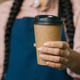 Close-up unrecognizable woman barista waitress in apron holding disposable paper cup with hot coffee - PhotoDune Item for Sale