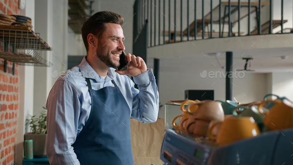 Caucasian man coffee shop employee in apron talk on mobile phone behind cafeteria counter - Stock Photo - Images