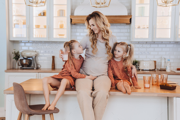 Pregnant Swedish mom sitting on kitchen table with little daughters dressed in beige casual clothes