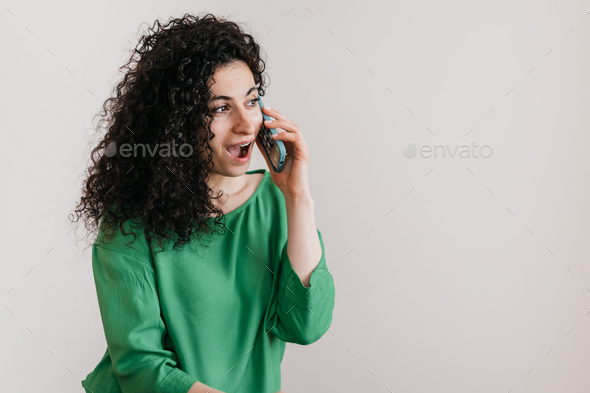 Excited Spanish young adult woman in green blouse talking by phone with opened mouth
