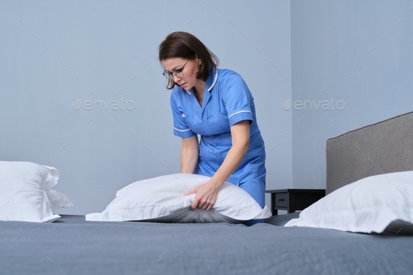 Middle aged female professional maid making bed in hotel room, laying out pillows