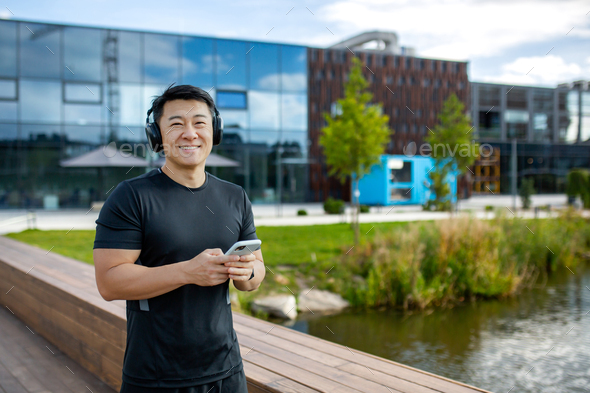 A young Asian male athlete in headphones and with a phone is standing outside and checking his - Stock Photo - Images