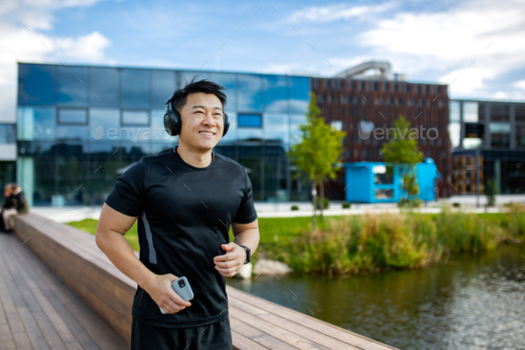 Smiling young Asian male athlete runner doing morning jog outside near lake wearing headphones and - Stock Photo - Images