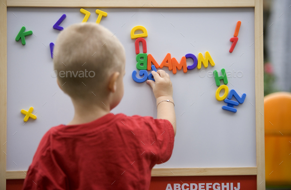 Back view of a blond toddler boy playing with colorful letters on a magnetic board - Stock Photo - Images