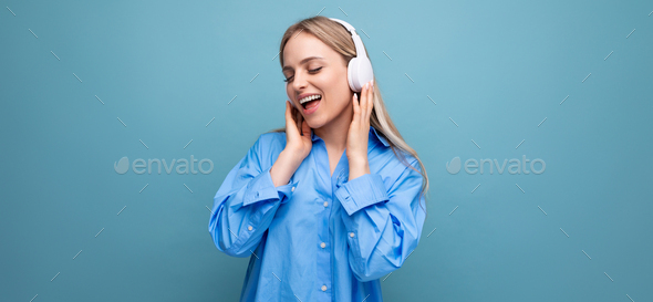 shy blonde girl in a casual shirt listens to songs in big headphones and sings along on a blue