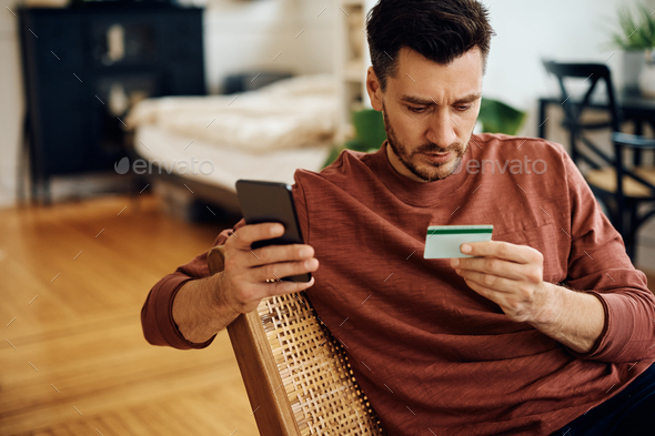Mid adult man using credit card and cell phone for online payment at home.