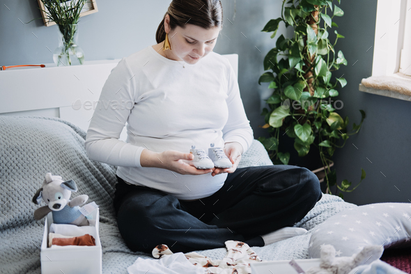 Pregnant woman with big belly holding bodysuit and sorting clothes for future. How to Organize