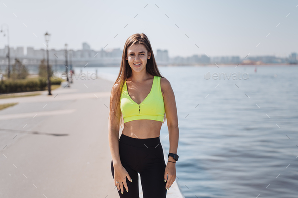 Stunning tanned Spanish girl in yellow sporty top and black sport leggings  standing at embankment Stock Photo by ionadidishvili