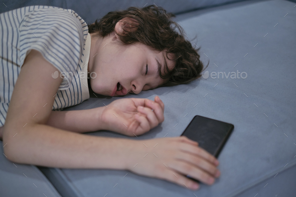 A 7-year-old boy sleeps on the couch at home with a phone in his hands, highlighting concerns about