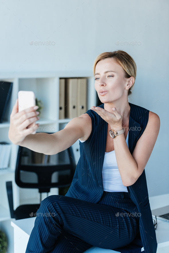 attractive adult businesswoman blowing air kiss during video call on smartphone in office