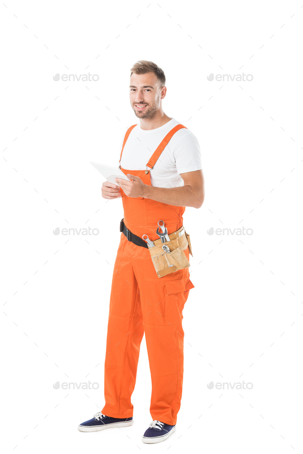 smiling handsome auto mechanic in orange uniform holding tablet isolated on white