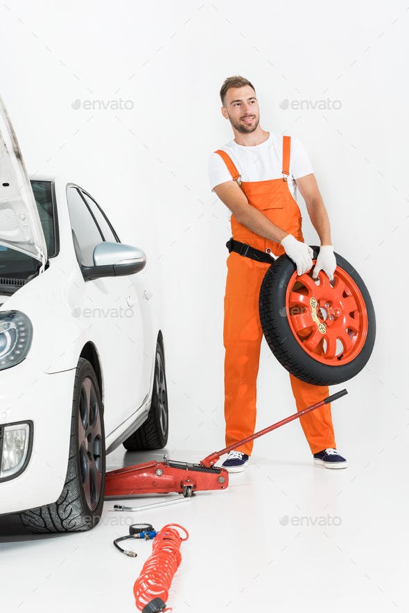 auto mechanic in orange uniform carrying car tire on white