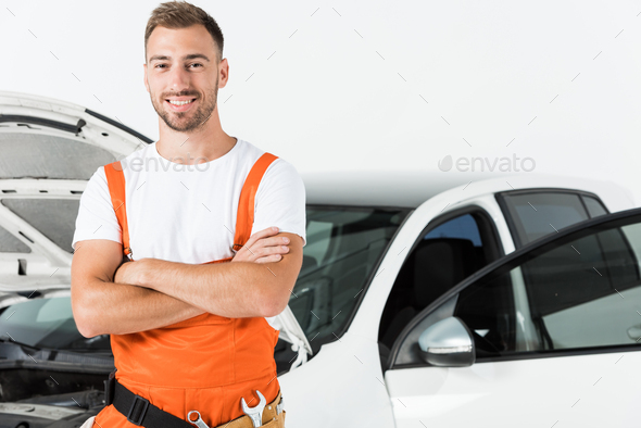 handsome auto mechanic in orange uniform standing with crossed arms near open car isolated on white
