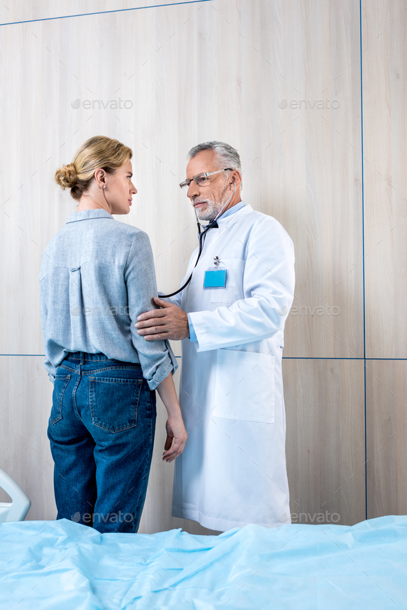 Confident Mature Male Doctor Examining Female Patient By Stethoscope In ...