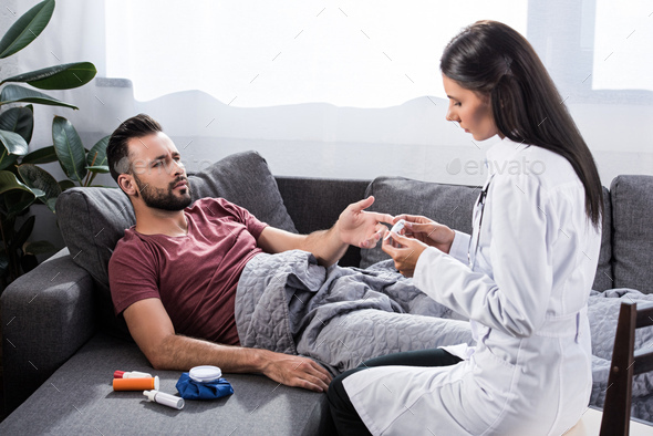 female doctor holding electric thermometer while her patient lying on couch and talking to her