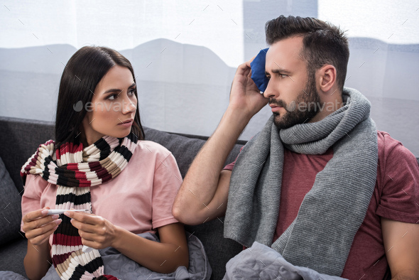 sick young couple with electric thermometer and ice pack sitting together on couch - Stock Photo - Images