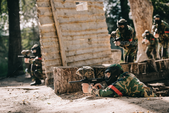 paintball player laying on ground and aiming by marker gun while his team hiding behind wooden wall