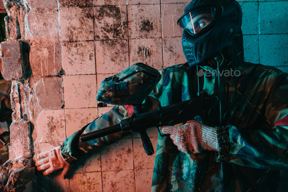 male paintball player in camouflage and goggle mask hiding behind wall and holding marker gun in