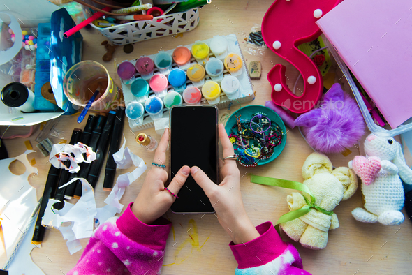 Hand of child girl using smartphone on chaotic desk in home office. Desk with objects for creativity