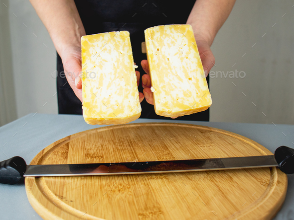 hands holding two pieces of marble cheese, cutting a head of cheese on a wooden cutting board