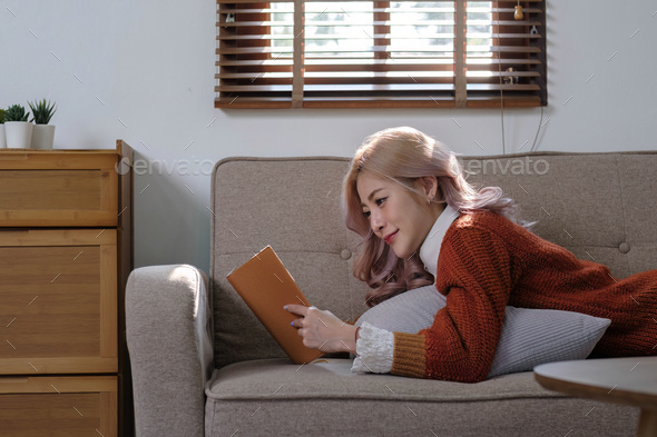 Attractive young Asian woman sits in the minimal and comfortable living room enjoying reading a book - Stock Photo - Images