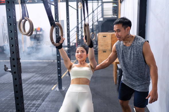 Male fitness instructor help woman doing pull-ups ring dips in a gym on gymnastic rings.