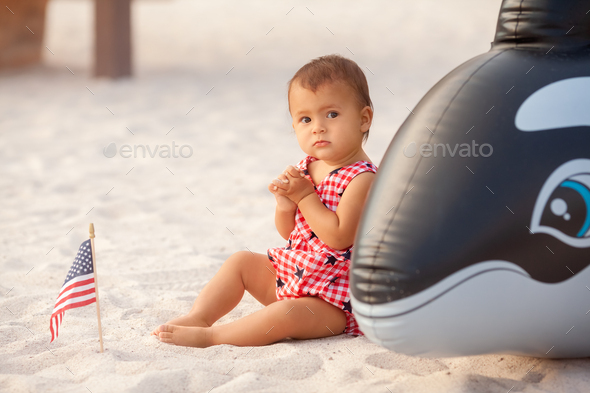 Infant baby girl in outfit of American flag sitting on sandy beach on 4th of July day - Stock Photo - Images