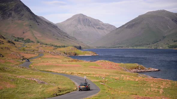 People driving on a road by the lake towards the mountains of the England Lake District - wide shot alt