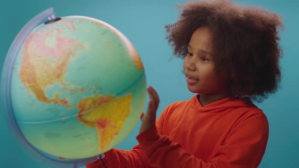 Curious African American school girl exploring the globe standing on blue background alt