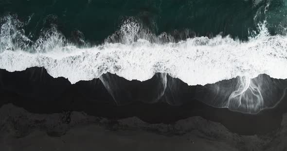 Overhead view of waves on a black sand beach with people walking on the shore alt