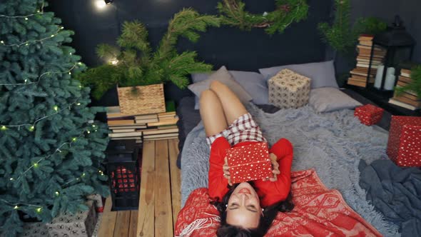 Pretty girl in cozy bedroom at Christmas. Beautiful young woman lying on bed with a gift in New year alt
