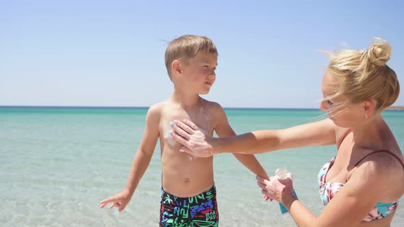 Cute Mother Puts Sunscreen Lotion on the Baby Azure Sea and Blue Sky in the Background Protection alt