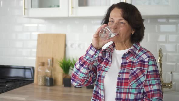 Happy Old Woman Drinking Fresh Water Standing in Home Kitchen. alt